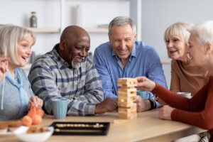 A senior adult smiles while unpacking a cardboard box in a cozy, sunlit apartment with family members nearby.